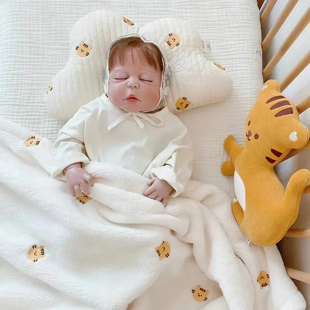 Une paisible bébé dort paisiblement dans un berceau douillet, blotti sous une couverture ornée de têtes de lion, avec une peluche lion et une Couverture de Bébé en Molleton de Corail de BABY PREMA pour l'hygiène à proximité.