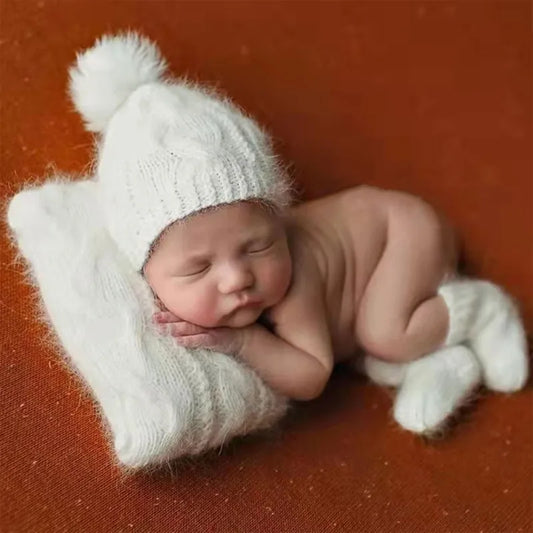 Un nouveau-né paisible dort paisiblement tout en portant un adorable bonnet tricoté blanc avec un pompon, complété par des mitaines blanches douillettes, le tout niché sur un fond chaud de couleur rouille fourni par l'ensemble Cadeau Naissance Bébé | 2 pièces pour Bébé.