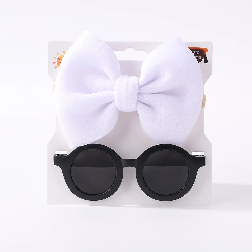 Un coffret contenant un Set d'Accessoires Bébé | Lunettes de Cheveux pour Bébé de BABY PREMA, comportant un grand nœud blanc et une paire de lunettes de soleil pour bébé rondes noires sur fond blanc.