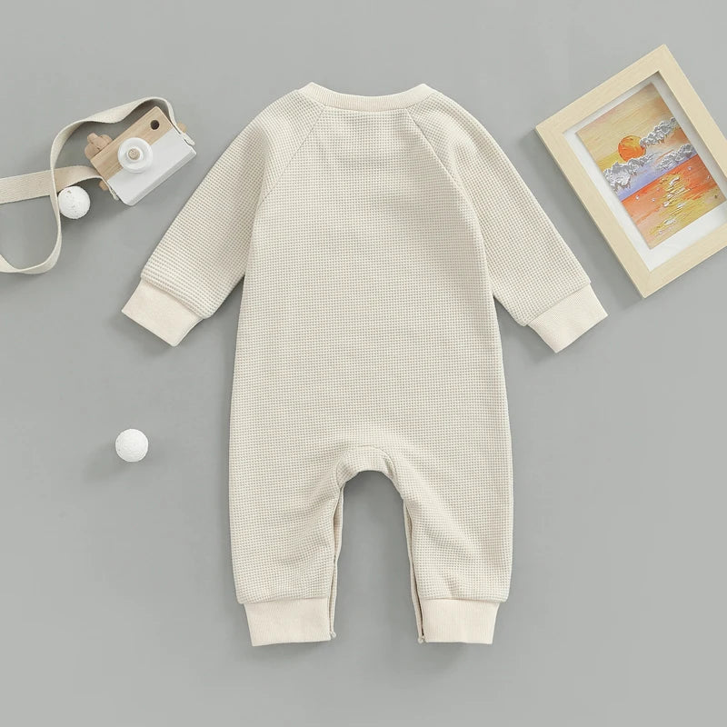 Genouillère Barboteuse | Naissance Bébé en beige avec manches longues et poignets côtelés, confectionné en tissu gaufré doux, présenté sur fond gris à côté d'un appareil photo jouet, d'un cadre photo et de deux petites boules de BABY PREMA.