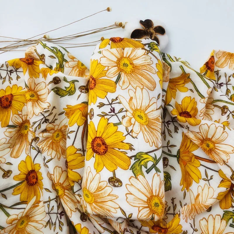 Un gros plan d'un tissu Lange Bébé en Bambou & Coton de BABY PREMA présentant un motif de marguerites jaunes avec des centres marron sur fond blanc, accentué par quelques plumes de dodo posées sur le dessus.