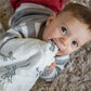 Un bébé curieux avec de grands yeux expressifs mordant de manière ludique un morceau de tissu doux et hygiénique tout en étant allongé sur une couverture en mousseline de coton pour bébé confortable et texturée "BABY PREMA".