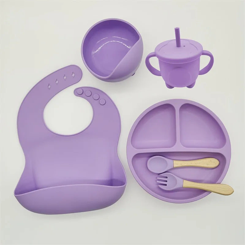 Un ensemble d'articles d'alimentation violets pour bébé, comprenant un bavoir, un bol, un gobelet, une assiette à trois sections, une cuillère et une fourchette en silicone de haute qualité, le tout soigneusement disposé sur une surface blanche. Le produit est l'Ensemble 6-8 Pièces de Vaisselle pour enfants de BABY PREMA.