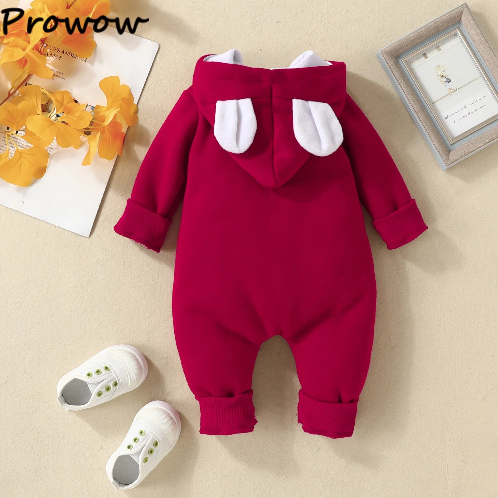 Une combinaison rouge pour bébé avec une capuche ornée de jolies oreilles blanches est disposée sur une surface beige. Cet adorable ensemble Combinaison à Capuche Pour Bébé comprend des chaussures de bébé blanches avec des boutons verts. A proximité, il y a une photo encadrée et un bouquet de fleurs jaunes. Le nom de la marque « BABY PREMA » est visible dans le coin supérieur gauche.