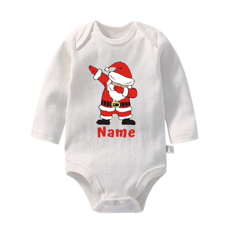 Une combinaison blanche pour bébé avec un graphisme du Père Noël faisant un pas de danse. Sous le graphique, le mot « Nom » est écrit, indiquant un espace pour la personnalisation. Ce Body Noël Personnalisé pour Bébé de BABY-PREMA est doté de manches longues et s'affiche sur un fond uni blanc.