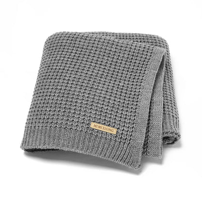 Une couverture en tricot gris pliée avec une petite étiquette beige qui dit "Baby-Prema", cette Couverture Bébé Tricot Fin 90*70cm - Baby-Prema est parfaite pour toutes les saisons. Fabriqué en acrylique doux, il assure confort et chaleur à votre tout-petit.