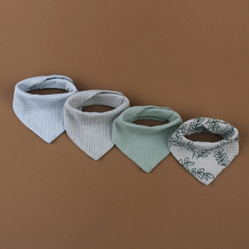 Quatre Bavoirs en mousseline de coton pour bébé de style bandana de BABY-PREMA sont disposés côte à côte sur une surface marron. Le lot de 4 bavoirs présente différentes couleurs et motifs : bleu clair, gris clair, vert et blanc avec un motif de feuilles vertes.