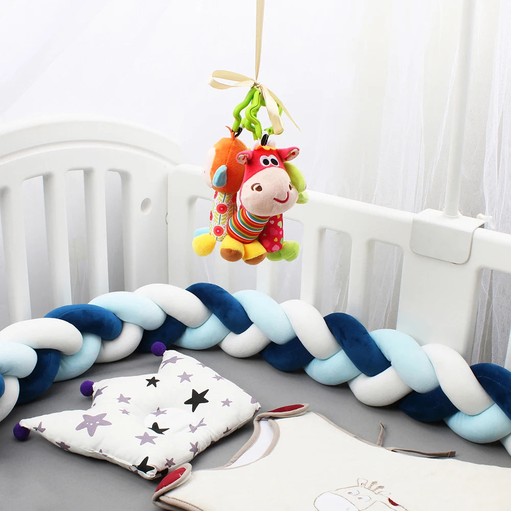 Un lit de bébé blanc est décoré d'un tour de lit tressé bleu et blanc. Au-dessus du berceau, une vache en peluche colorée est suspendue à un ruban. À l'intérieur du berceau, il y a un oreiller à motif étoilé et une tenue de bébé beige sans manches avec un motif animal brodé. Cette configuration élégante comprend le Tour de Lit Tressé Noué de BABY PREMA pour plus de confort.