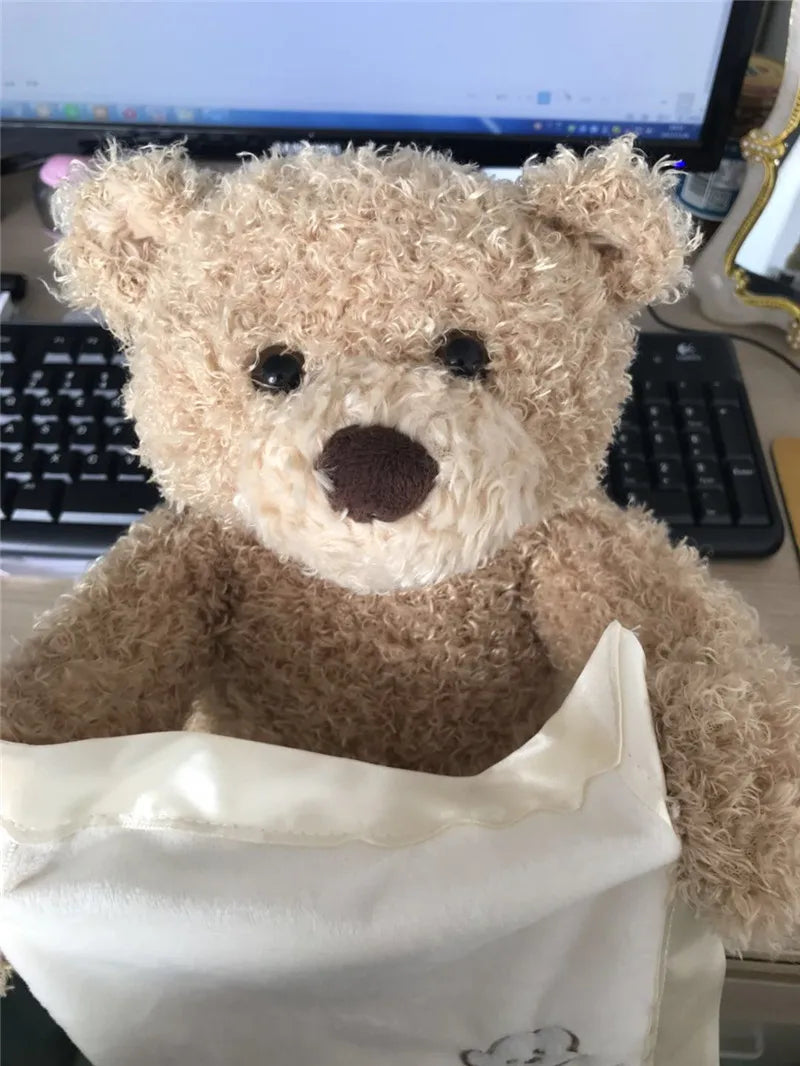 Un Doudou Ours Peluche Musical Chantant "Peekaboo" sortant d'un sac en tissu de couleur crème, posé sur un bureau avec un clavier d'ordinateur en arrière-plan, destiné à un enfant prématuré. (Nom de marque : BÉBÉ PREMA)