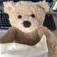 Un doudou peluche Teddy Ours Musical "Peekaboo" de BABY PREMA qui sort d'un sac en tissu de couleur crème sur un bureau avec un clavier d'ordinateur en arrière-plan, prêt à dodo.