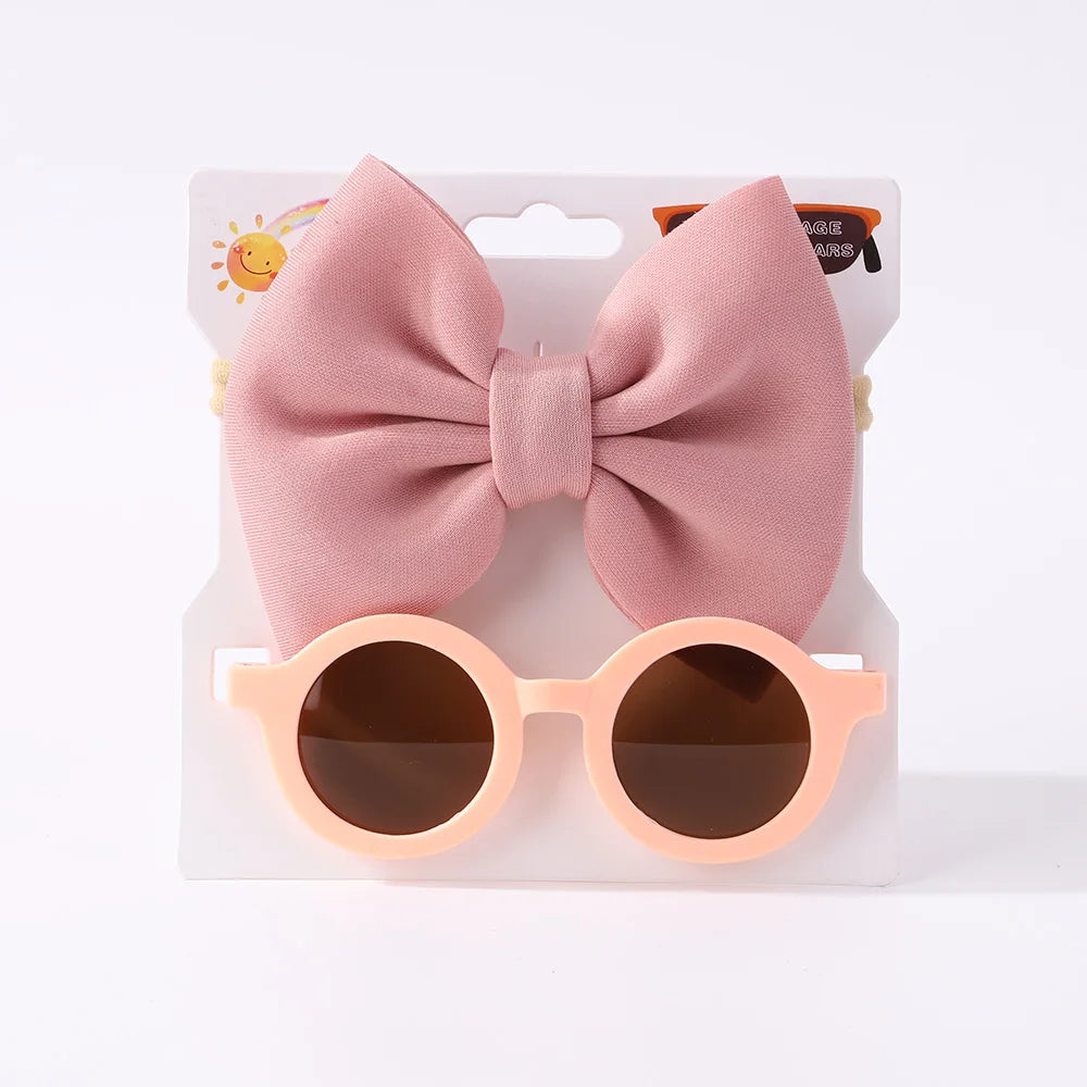 Une paire de lunettes de soleil pour bébé rondes rose clair avec des verres foncés et un grand nœud rose clair sont présentées sur un fond blanc. Le nom du produit est Set d'Accessoires Bébé | Lunettes de Cheveux pour Bébé de la marque BABY PREMA.