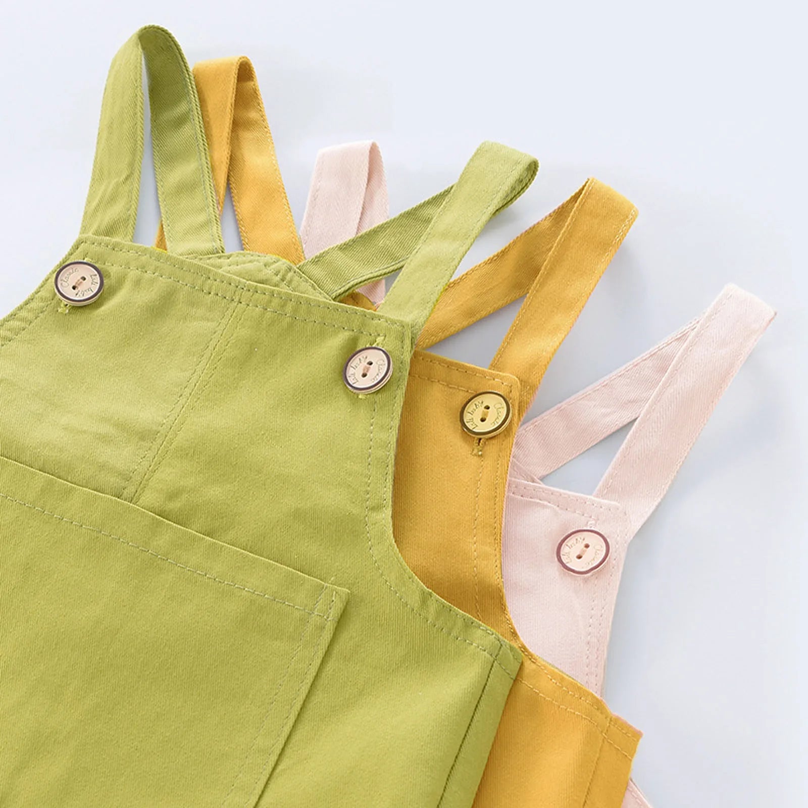 Trois Salopette Bébé Unie de BABY-PREMA en vert, jaune et beige sont empilées, mettant en valeur leurs charmants détails de boutons sur les bretelles. Ces ravissantes options de vêtements pour enfants sont disponibles en coton doux ou en denim résistant.