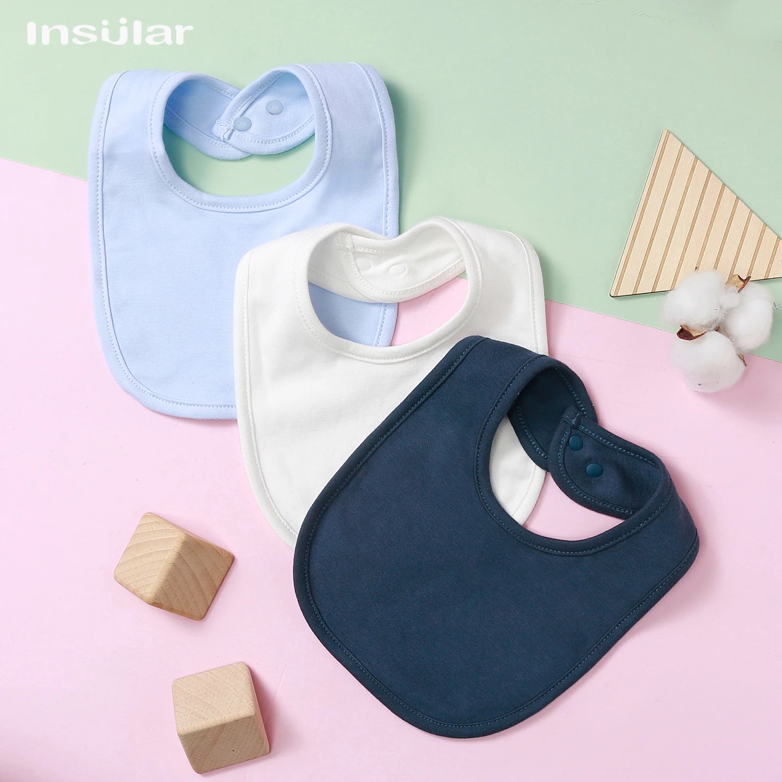 Les essentiels de bébé dans un agencement minimaliste : des bavoirs BABY PREMA Bavoir Bébé en Coton Bio bleu pastel, blanc et marine associés à des blocs de bois et un brin de coton sur un bicolore rose et bleu.