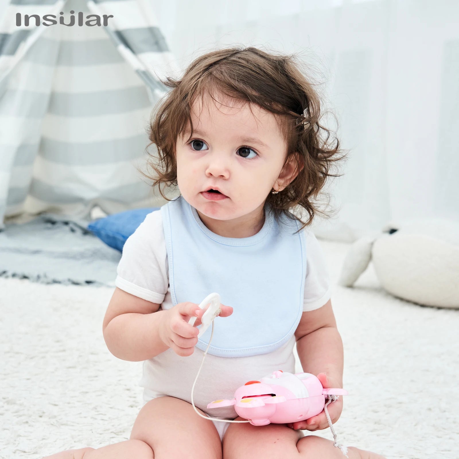 Bambin curieux avec un jouet, vivant une récréation sur un tapis douillet et entouré des articles Bavoir Bébé en Coton Bio de BABY PREMA.