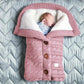 Un nouveau-né paisible dort confortablement dans une gigoteuse tricotée rose en forme de cosse de pois, dotée de boutons en bois et d'accessoires essentiels pour bébé, la Couverture de Poussette Pour Bébé de BABY PREMA.