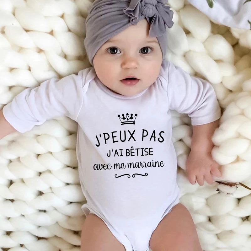 Un bébé avec un bandeau gris et une barboteuse en coton blanc repose sur une douce couverture blanche. La barboteuse comporte le texte français qui dit : « J'peux pas, j'ai bêtise avec ma marraine », qui se traduit par « Je ne peux pas, je me fais des ennuis avec ma marraine ». Parfait comme cadeau pour nouveau-né ! Le Body coton Manches Longues de BABY-PREMA est un choix idéal pour cette adorable tenue.