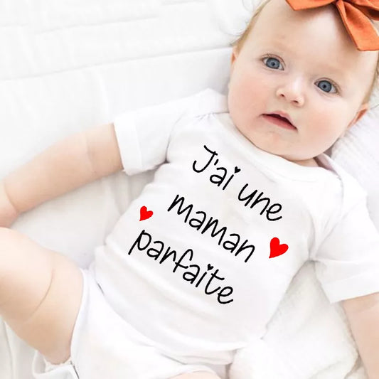 Un bébé est allongé sur une surface blanche et porte un Body Bébé Coton Imprimé Préma blanc avec le texte français "J'ai une maman parfaite" et deux illustrations de cœurs rouges. Le bébé, aux cheveux clairs et avec un noeud orange sur la tête, met en valeur une charmante pièce du garde-robe enfant BABY-PREMA.