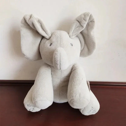 Un jouet Doudou Eléphant Peluche Musicale de BABY-PREMA assis contre un mur blanc sur une surface plane, avec une douce fourrure grise et des oreilles tombantes, dégageant un aspect réconfortant et câlin pour bébé.