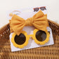 Une paire de lunettes de soleil pour bébé en forme de fleur jaune et des bandeaux à nœuds en nylon assortis de l'ensemble d'accessoires bébé BABY PREMA | Les Lunettes de Cheveux pour Bébé sont présentées sur un support cartonné, placé dans un panier en osier.