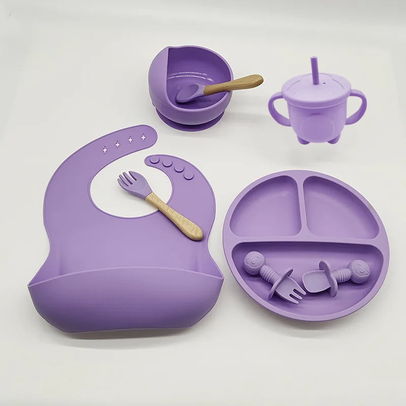 Un ensemble d'articles d'alimentation pour bébé violets en silicone de haute qualité, comprenant un bavoir, un bol avec une cuillère, un gobelet et une assiette divisée avec des ustensiles. Chaque article est soigneusement conçu pour les tout-petits et placé sur un fond blanc est l'Ensemble 6-8 Pièces de Vaisselle pour enfants de BABY PREMA.