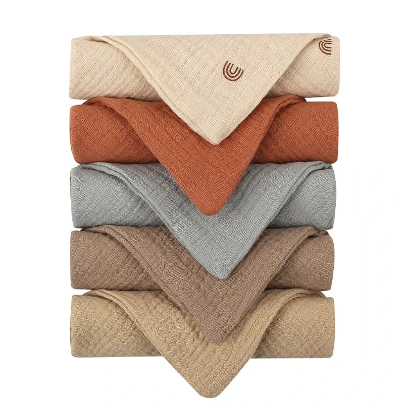 Un assortiment soigneusement empilé de Lot de 5 Langes Bébé en Coton pliés dans différents tons terreux, notamment beige, rouille, gris et taupe, avec un emblème dodo brodé visible sur la serviette supérieure de BABY PREMA.