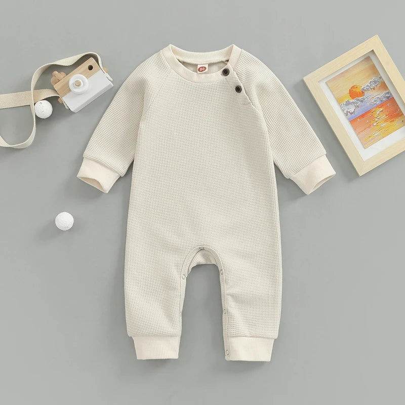 Une Genouillère Barboteuse beige | Naissance Bébé avec boutons latéraux sur fond gris, accompagné d'un appareil photo jouet, de deux boules blanches et d'un tableau encadré d'un coucher de soleil de BABY PREMA.