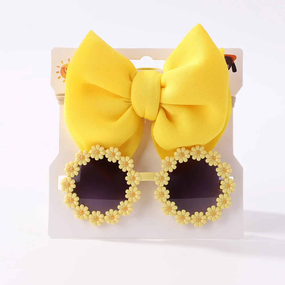 Un grand nœud jaune et une paire de lunettes de soleil rondes avec une monture en forme de fleur jaune en forme de marguerite, parfaites comme Set d'Accessoires Bébé | Les Lunettes de Cheveux pour Bébé de BABY PREMA, s'affichent sur un fond blanc.