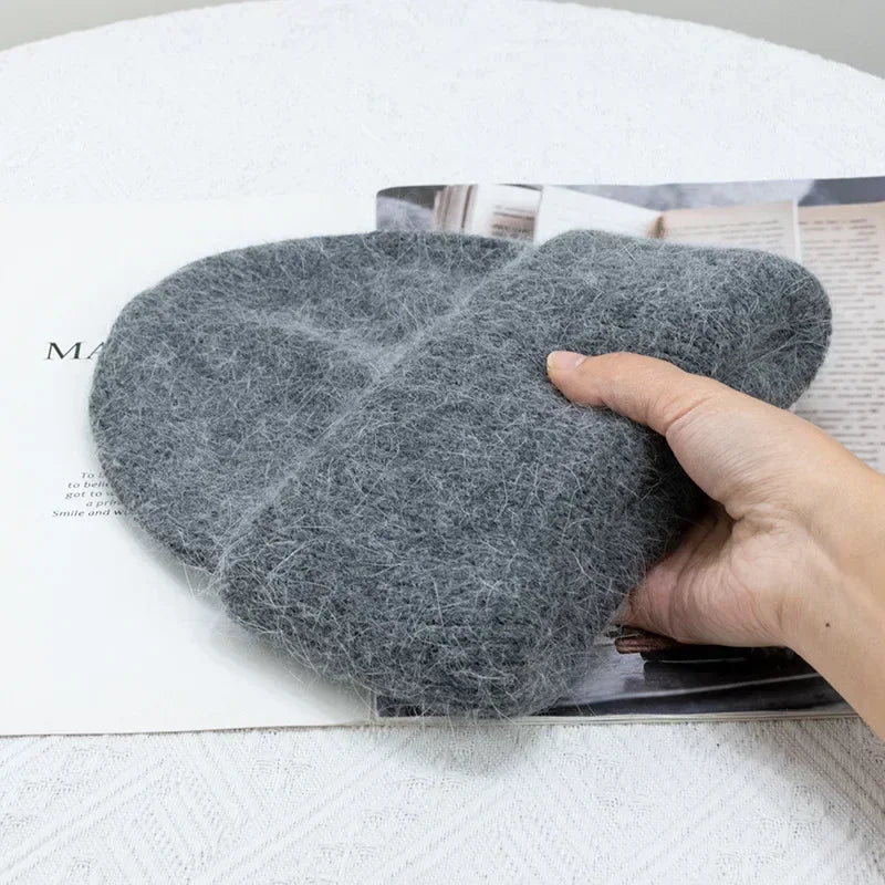 Une main tenant une paire de chaussons douillets en laine feutrée grise au-dessus d'un magazine ouvert, présentant un mélange d'artisanat fait main et d'accessoires de loisirs pour bébés, dont le Bonnet Angora Pour Maman de BABY PREMA.