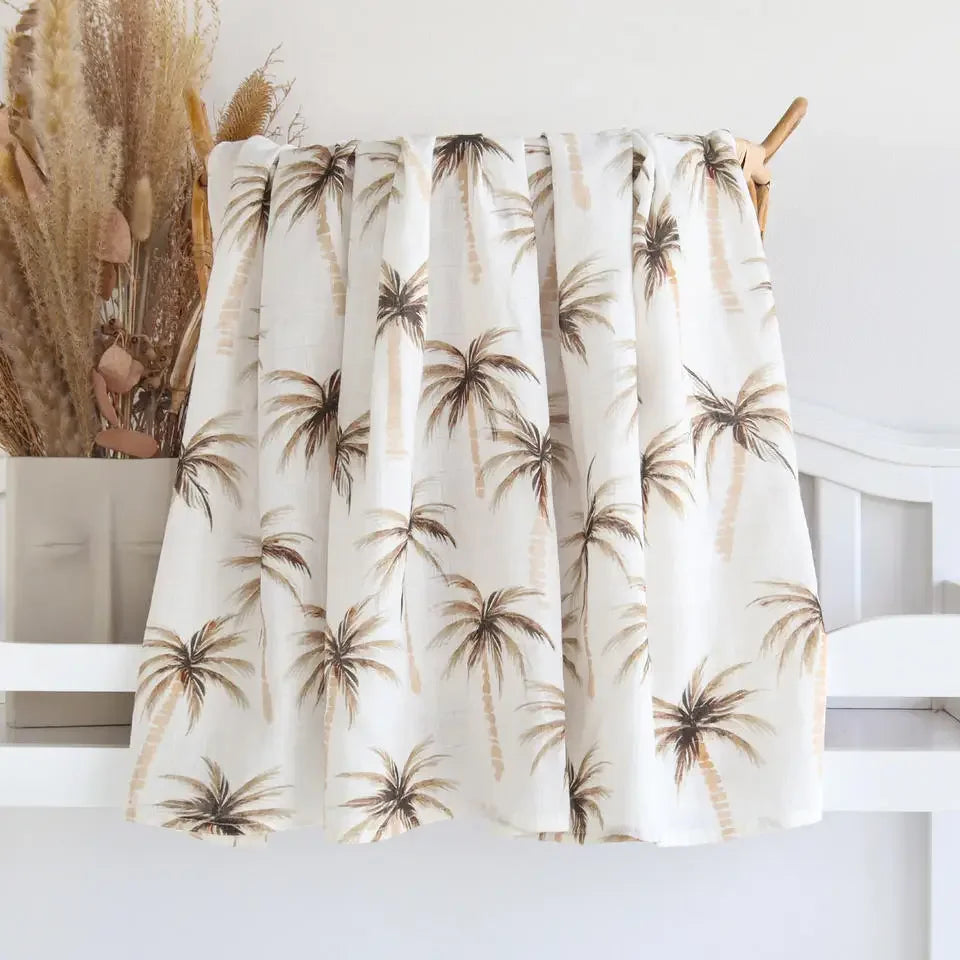 A beige Couvertures en Bambou Bébé | Emmaillotage à imprimé palmier tropical accroché sur un cintre en bois sur fond neutre avec plantes séchées décoratives par BABY PREMA.