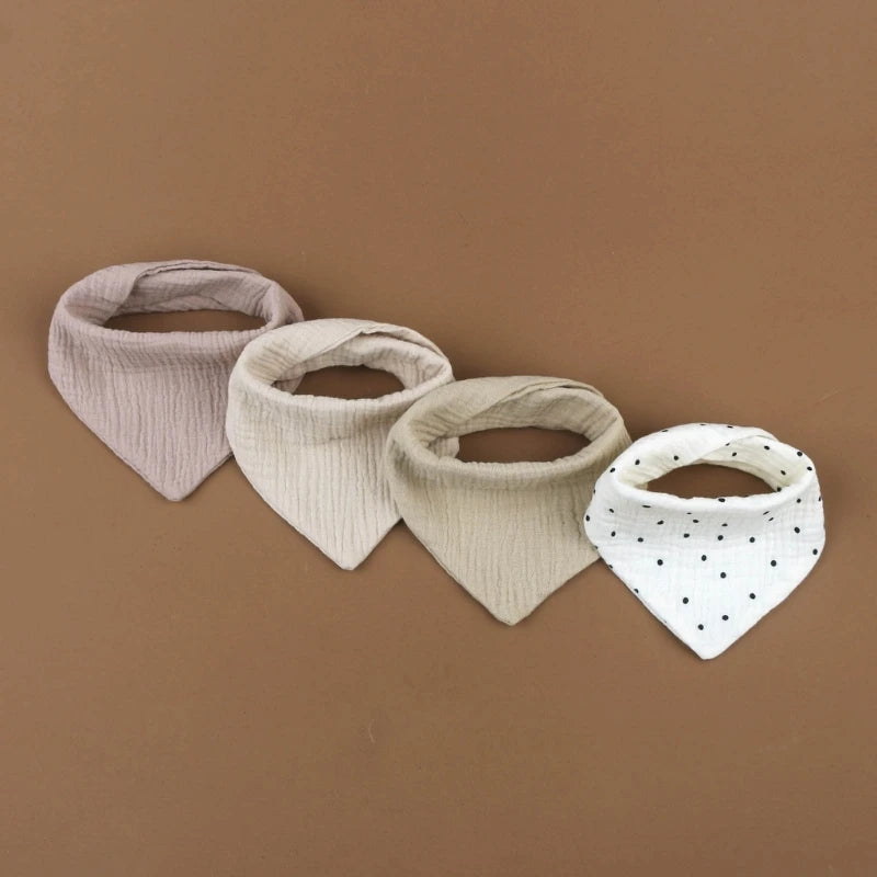 Quatre BABY-PREMA Bavoirs en mousseline de coton pour bébé sont disposés en rangée sur un fond marron. De gauche à droite, les bavoirs en mousseline de coton sont rose uni, gris clair, beige et blanc à pois noirs. Chaque bavoir a la forme d'un bandana et est fabriqué dans un tissu doux et froissé.