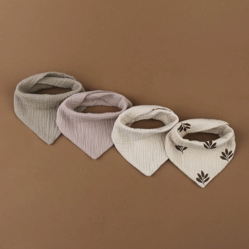 Quatre bavoirs en coton doux sont disposés côte à côte sur une surface marron. Ce Bavoirs en mousseline coton pour bébé de BABY-PREMA affiche différentes couleurs sourdes : la première est grise, la seconde est un lilas clair, la troisième est blanche et la quatrième est également blanche avec des petits motifs de feuilles marron.