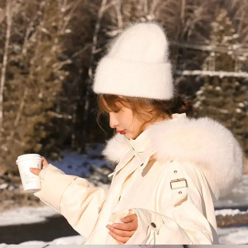 Une personne en tenue d'hiver, avec un bonnet Angora Pour Maman douillet et un manteau doublé de fourrure conçu comme accessoires bébé, profitant de la chaleur d'une boisson chaude par une journée ensoleillée et enneigée.