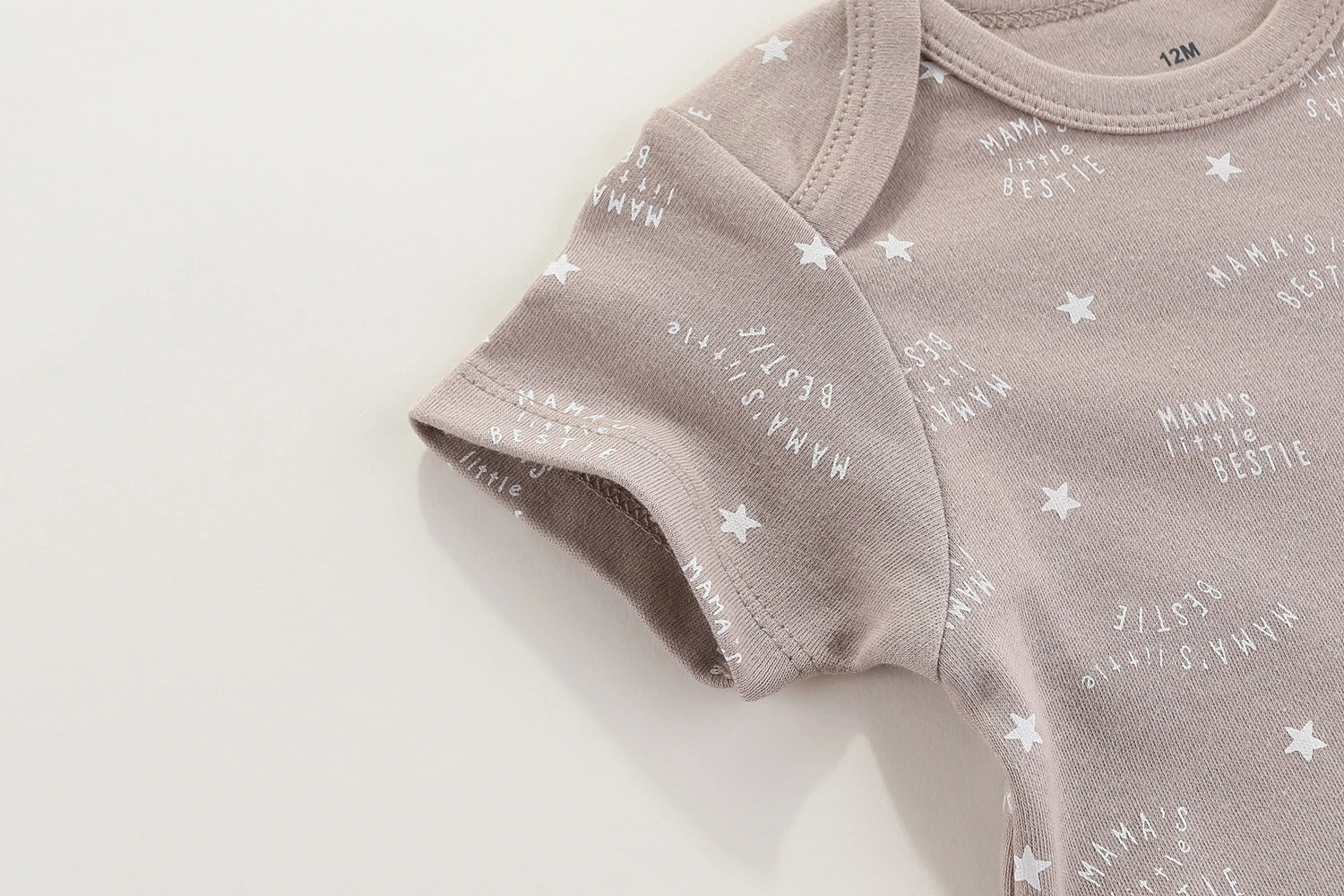 Gros plan d'une combinaison beige pour bébé avec des étoiles blanches et un texte indiquant « La petite meilleure amie de Mama » imprimé à plusieurs reprises. Confectionnée à partir de bodys doux en coton par BABY PREMA, cette adorable pièce présente des manches courtes et une étiquette de taille indiquant 12 mois. Le nom du produit est Lot 6 Body Coton Bébé Manches Courtes.