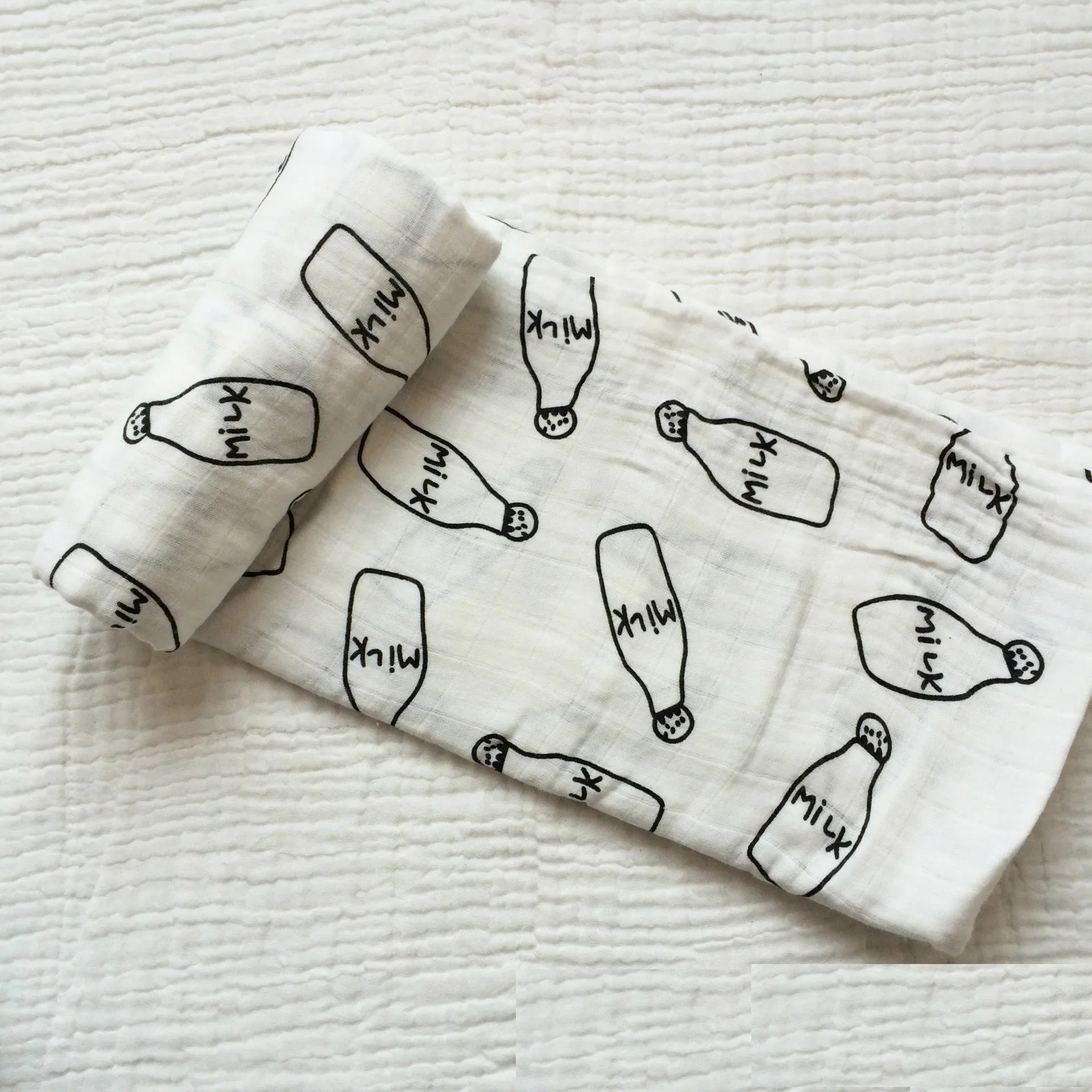 Une couverture en mousseline de coton blanc avec de mignonnes illustrations de bouteilles de lait et de sucettes glacées éparpillées sur sa surface, parfaite comme accessoire pour bébé BABY PREMA.