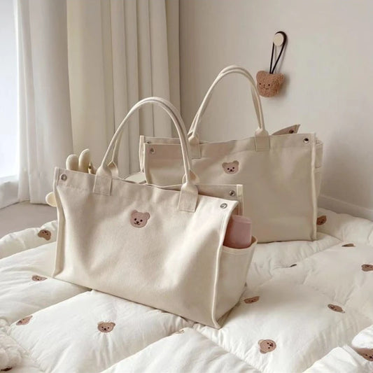 Deux cabas Sac à Langer Bébé Fourre Tout Beige de BABY-PREMA, avec de jolis visages d'ours, posés sur une douce couverture matelassée aux motifs d'ours assortis, évoquent une sensation de style douillet et minimaliste, parfaits pour transporter les essentiels de mon bébé.