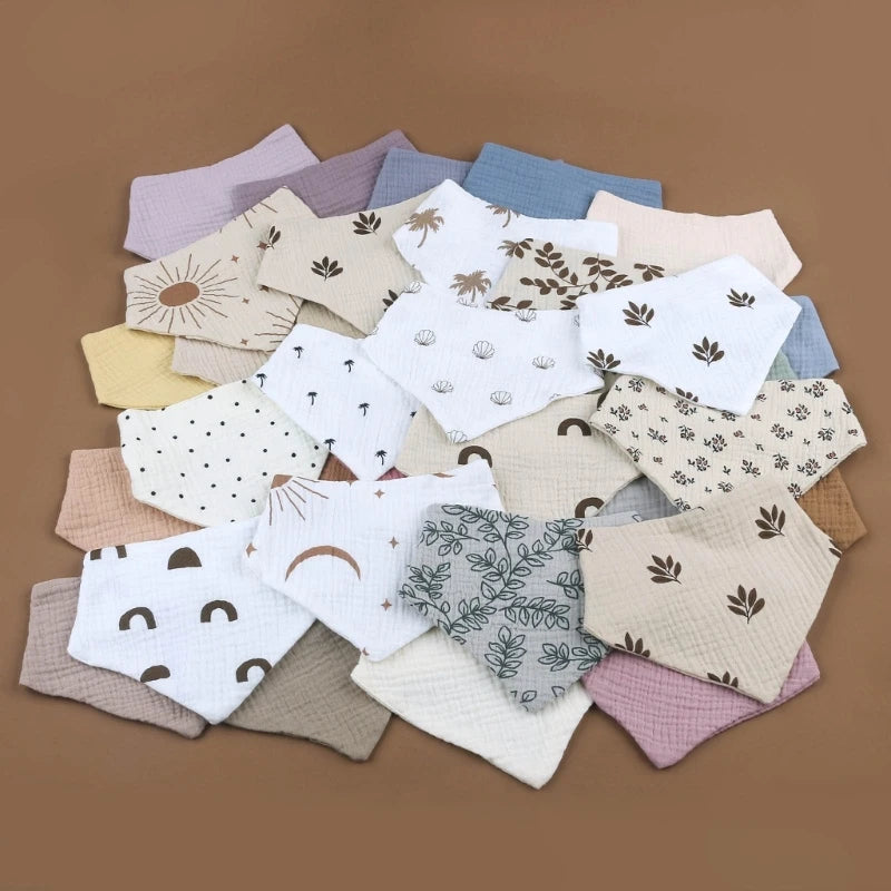 Une collection de bavoirs bandana pour bébé aux motifs et couleurs variés, soigneusement disposés sur une surface marron. Ce Bavoirs en mousseline de coton pour bébé présente des motifs tels que des arcs-en-ciel, des feuilles et des formes abstraites dans des tons pastel et neutres, dont le blanc, le beige, le bleu clair et le mauve. Fabriqué en mousseline de coton douce par BABY-PREMA.