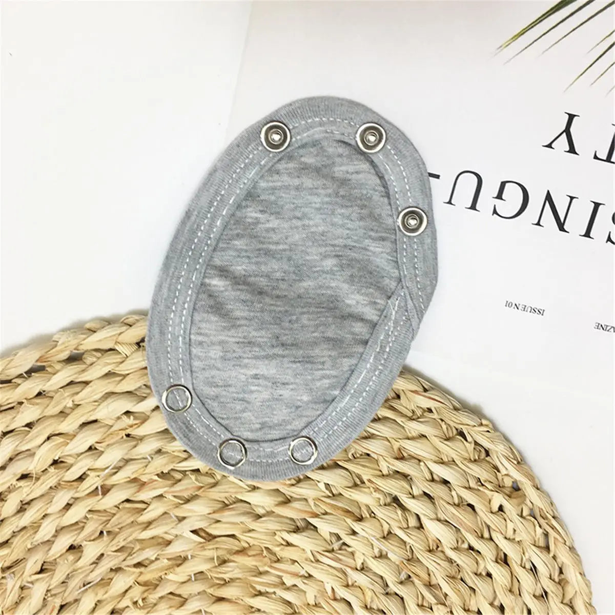 Coussinet de forme ovale en tissu gris avec boutons pression sur fond paille tressée, idéal pour l'Extenseur de Body Bébé 100% Coton BABY-PREMA.