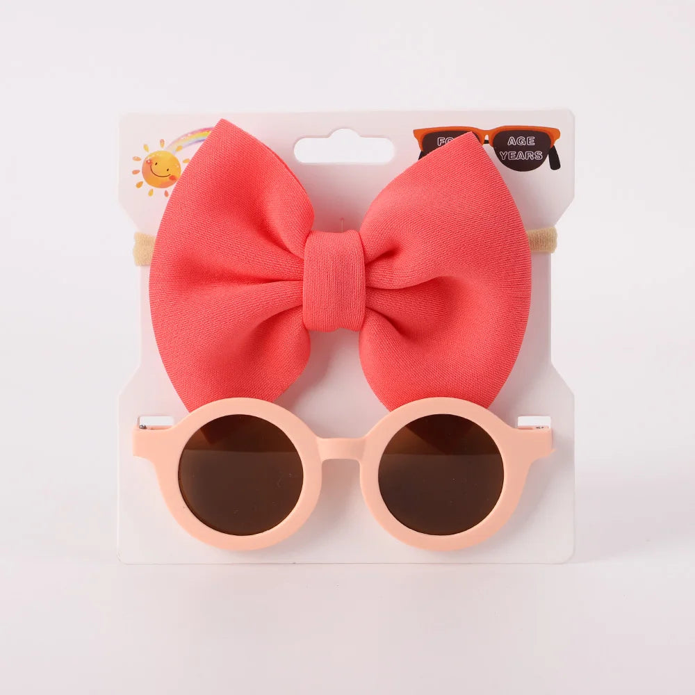 A BABY PREMA Ensemble d'Accessoires Bébé | Les lunettes de Cheveux pour Bébé avec des verres foncés ronds à monture rose et un grand bandeau à nœud en nylon rose sont présentées sur un support en carton blanc, avec un petit graphique solaire dans le coin supérieur.