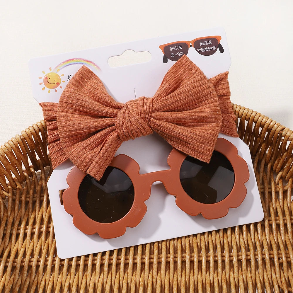 Une paire de lunettes de soleil pour bébé marron en forme de fleur et un nœud pour cheveux marron assorti affichés sur une carte blanche, placés dans un panier en osier de l'ensemble d'accessoires bébé | Lunettes de Cheveux pour Bébé de BABY PREMA.