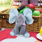 Jouet éléphant en peluche Baby-Prema assis devant un panier de pique-nique avec des œufs colorés par une journée ensoleillée, apparemment prêt pour une joyeuse célébration de Pâques conçue pour un enfant prématuré.