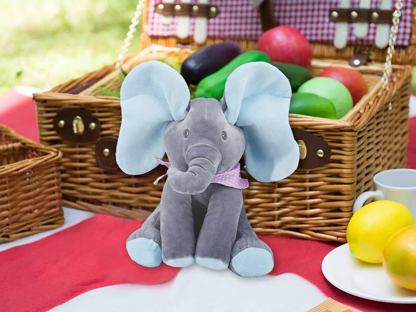 Jouet éléphant en peluche Baby-Prema assis devant un panier de pique-nique avec des œufs colorés par une journée ensoleillée, apparemment prêt pour une joyeuse célébration de Pâques conçue pour un enfant prématuré.