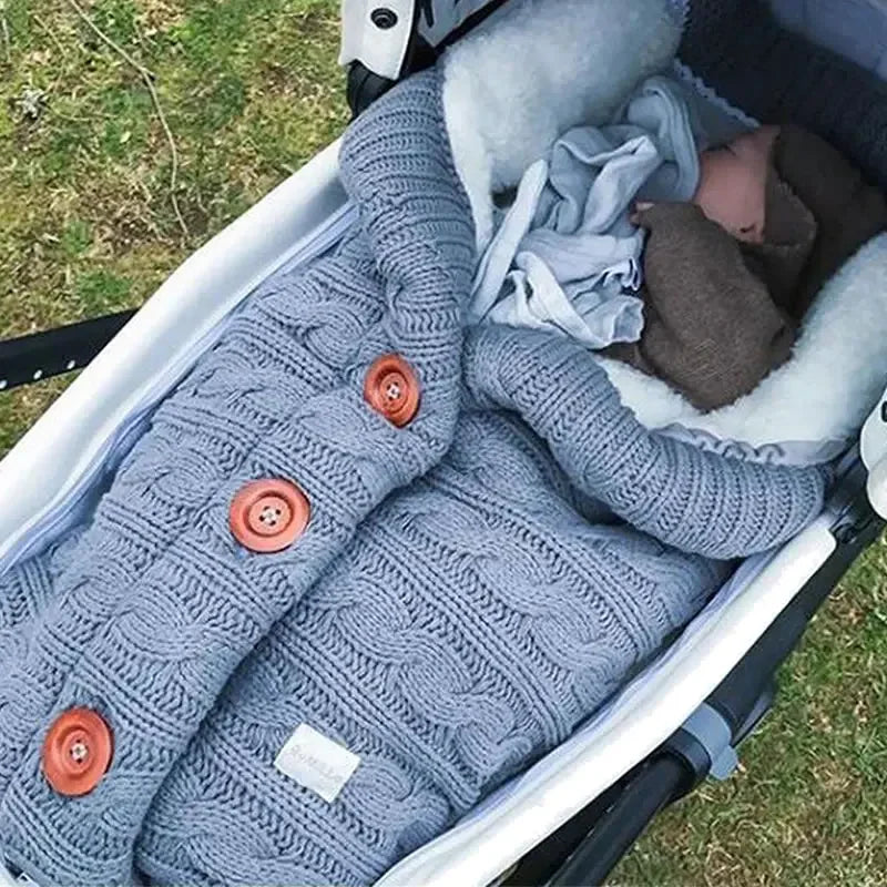 Un bébé confortablement enveloppé dans une couverture de poussette pour bébé douillette en tricot torsadé avec de gros boutons orange, endormi paisiblement à l'extérieur parmi leurs Accessoires bébé de BABY PREMA.