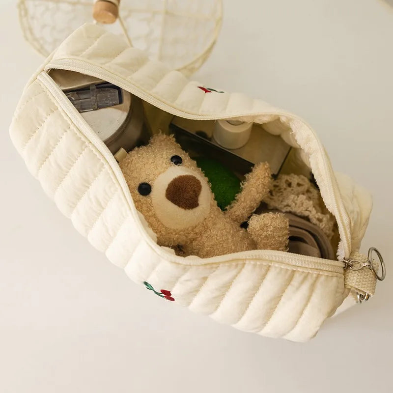 Un adorable ours en peluche sort d'une pochette en tissu matelassé de couleur crème remplie de quelques petits objets, prêt pour une aventure avec un BABY PREMA Sac à Couches en Coton pour poussette.