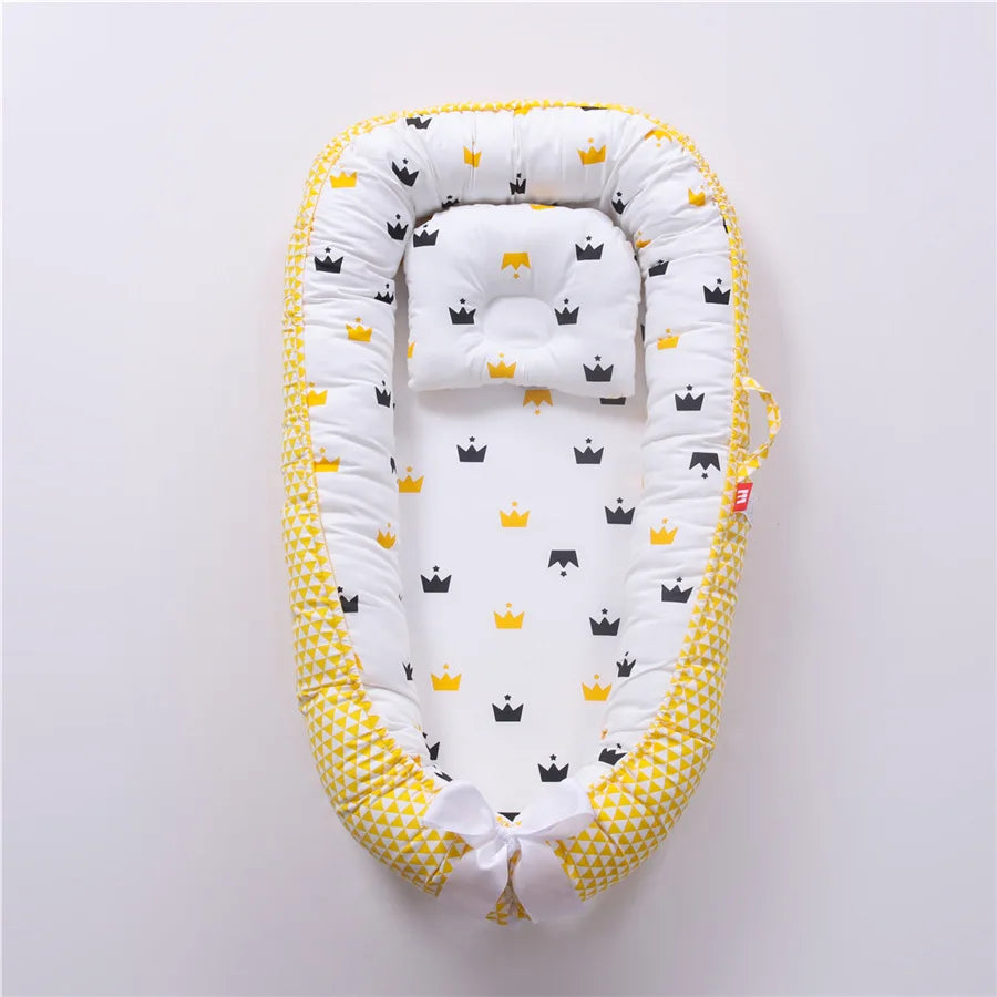 Un Lit de Bébé portable Nid de Cocoon pour bébé avec un design blanc et jaune, avec de jolies couronnes noires sur fond blanc, offrant un espace confortable et sécurisé pour qu'un enfant se repose par BABY-PREMA.