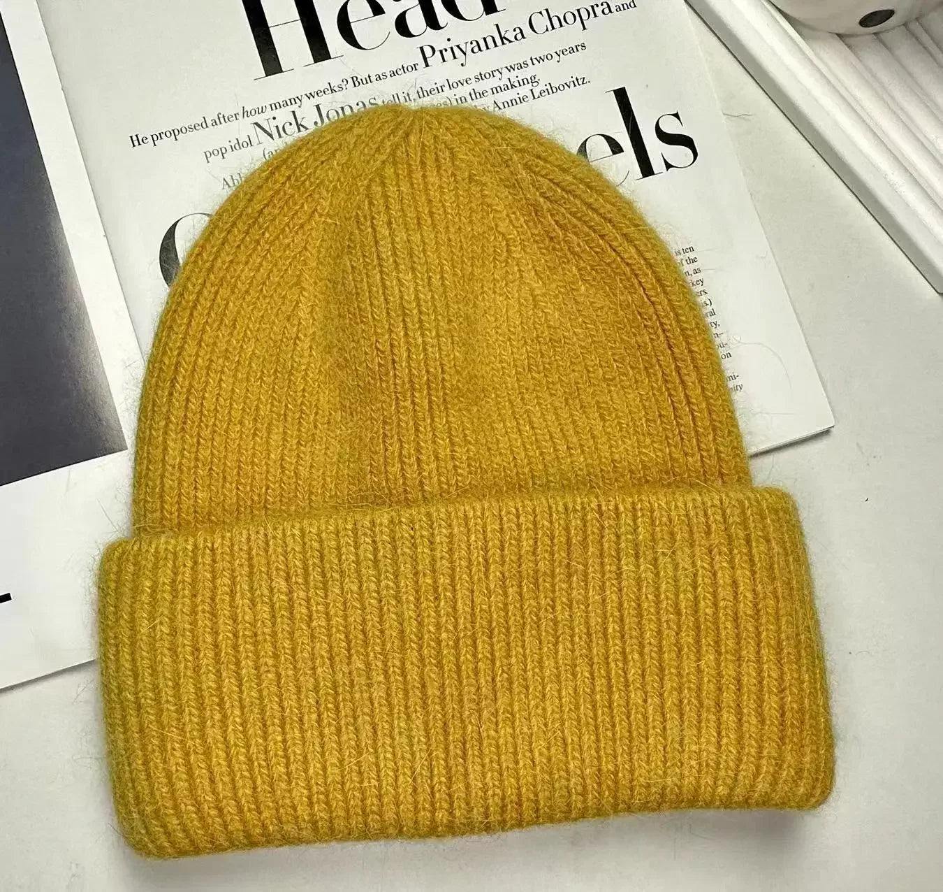 Un bonnet pour bébé Bonnet Angora Pour Maman jaune moutarde de BABY PREMA repose sur un journal ouvert, ajoutant une touche de couleur à l'imprimé noir et blanc.