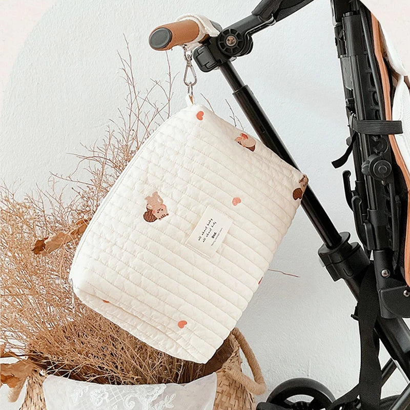 Un élégant Sac à Couches en Coton pour poussette de BABY PREMA avec de jolis motifs d'ours accrochés au guidon d'un scooter plié, sur un fond minimaliste avec des plantes séchées et une étiquette « mon bébé » bien en évidence.