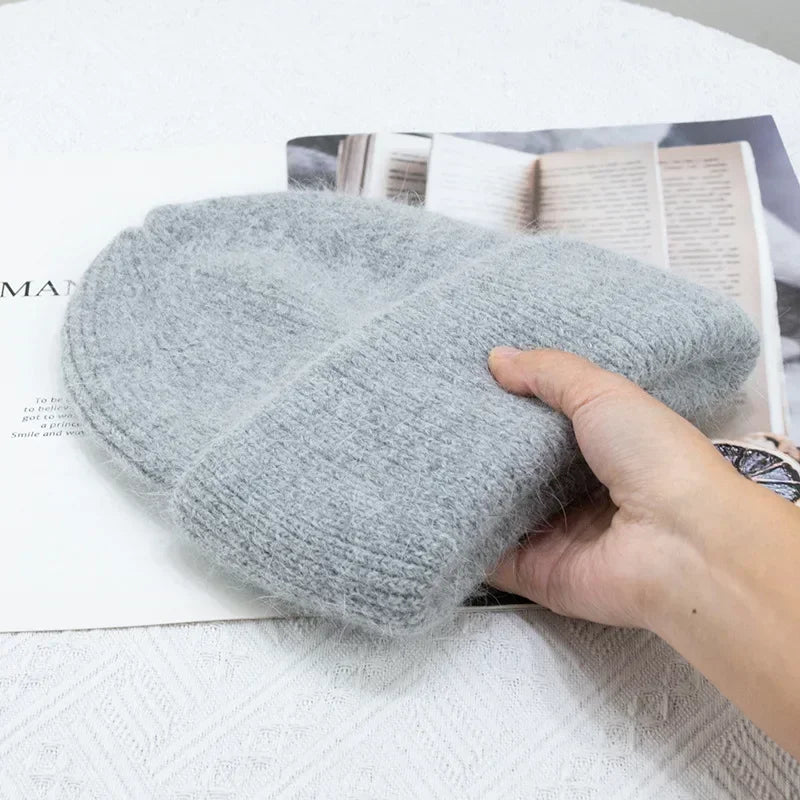 Une confortable paire de mitaines tricotées grises et moelleuses posées sur un magazine ouvert, avec la main d'une personne qui les tient doucement, illustrant un accessoire confortable pour prendre soin du Bonnet Angora Pour Maman de BABY PREMA.