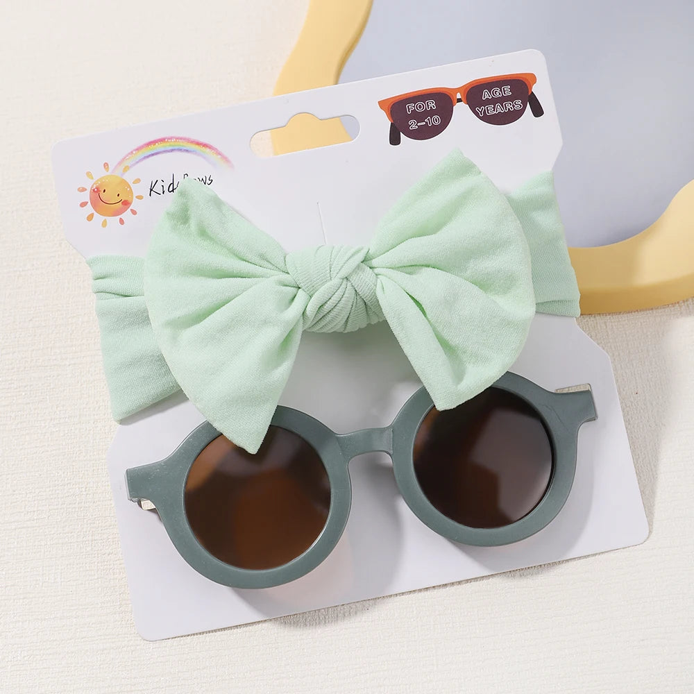 Une paire de lunettes de soleil pour bébé rondes grises et un bandeau vert menthe avec un grand nœud sont attachés à une carte d'emballage blanche étiquetée « Set d'Accessoires Bébé | Lunettes de Cheveux pour Bébé by BABY PREMA.