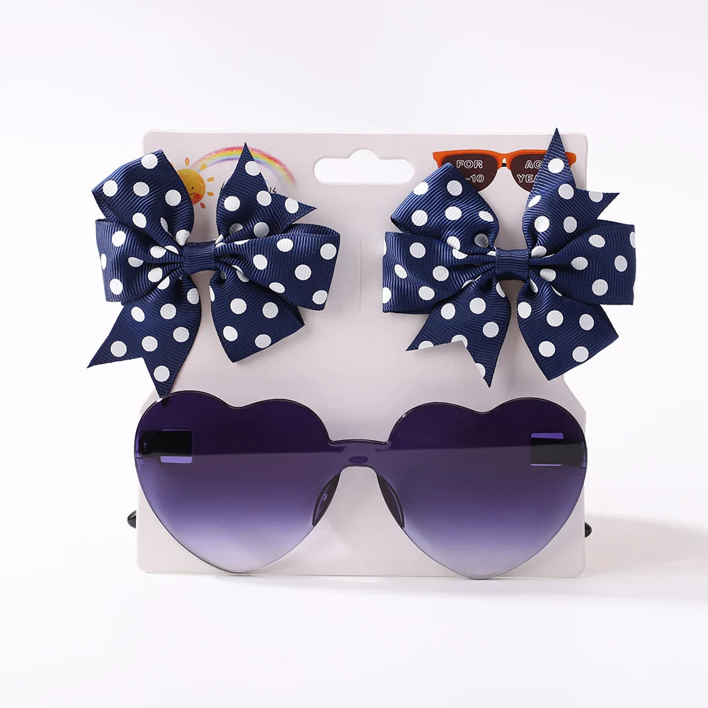 A **Ensemble d'Accessoires Bébé | Lunettes de Cheveux pour Bébé** de **BABY PREMA** avec de grandes lunettes de soleil en forme de cœur avec des verres foncés et deux nœuds pour cheveux à pois bleus attachés à la carte d'affichage, parfaits pour ajouter une touche de style à tous les accessoires pour cheveux de bébé.