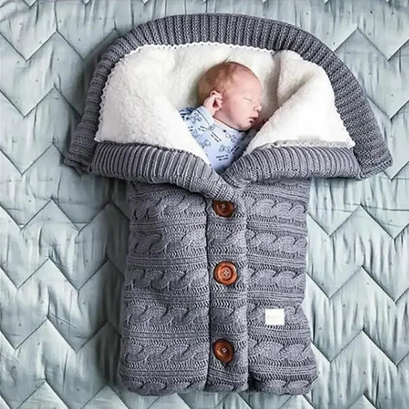 Un nouveau-né paisible dort confortablement enveloppé dans un sac douillet en forme de pull qui ressemble à un sac de couchage avec des détails de boutons, sur un fond gris à motifs. Cet article est un complément parfait à
Couverture de Poussette Pour Bébé de BABY PREMA.
